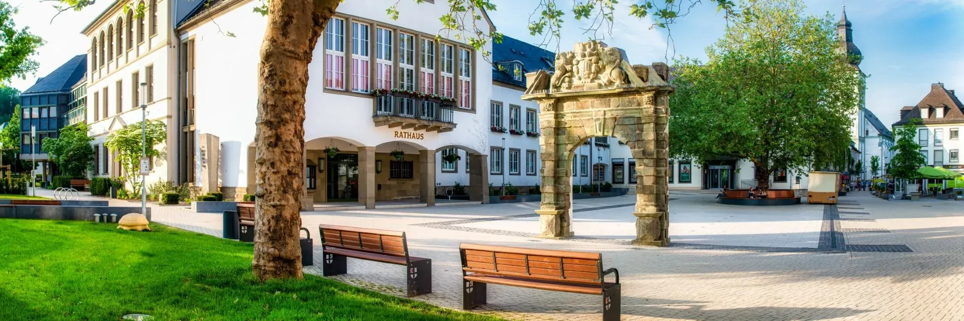 Sonniger Rathausplatz mit historischem Steinportal, Rathaus im Hintergrund und Bänken unter Bäumen.